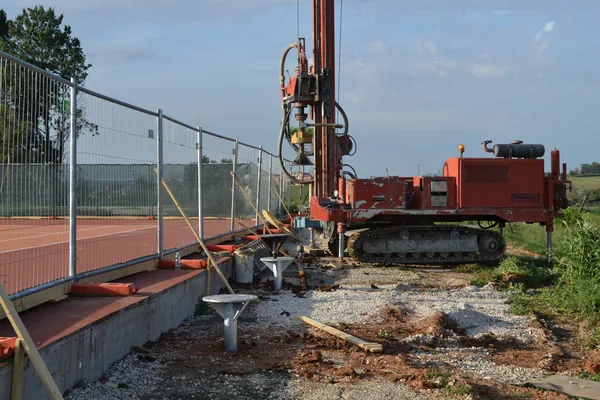 Bordo di un campo da tennis in terra rossa: fondazioni a vite T-Block già posate lungo la recinzione e la trivella cingolata al lavoro.