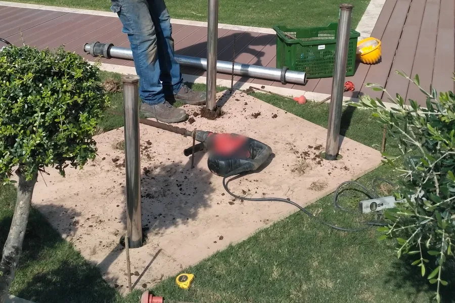 Posa nel giardino di un resort: quattro fondazioni a vite T-Block già avvitate nell'aiuola attorno a un albero, con l'avvitatore elettrico appoggiato a terra accanto.