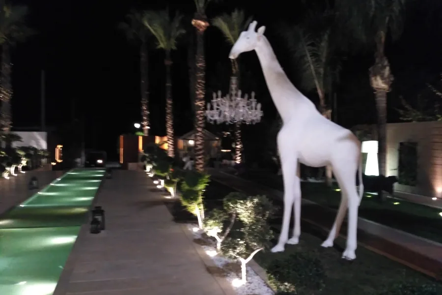 Una scultura Giraffe in Love illuminata di notte tra le palme del parco di un resort a Paestum, con i lampadari di cristallo accesi lungo il viale.