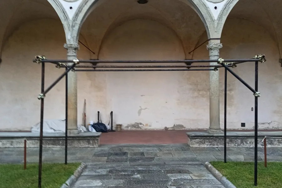Telaio in tubo da ponteggio montato sul prato del Secondo Chiostro di Santa Croce, davanti a due arcate del loggiato rinascimentale con i tondi in pietra.