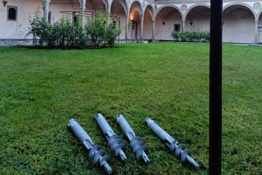 Quattro fondazioni a vite T-Block zincate appoggiate sul prato del Secondo Chiostro di Santa Croce, pronte per la posa, con il loggiato ad archi sullo sfondo.