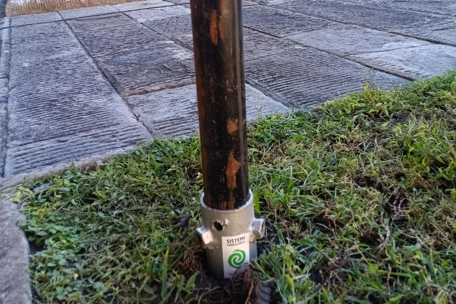 T-Block senza testa per palo Ø48 a filo del prato, con il tubo da ponteggio infilato e i dadi laterali pronti per il bloccaggio; dietro, il chiostro.