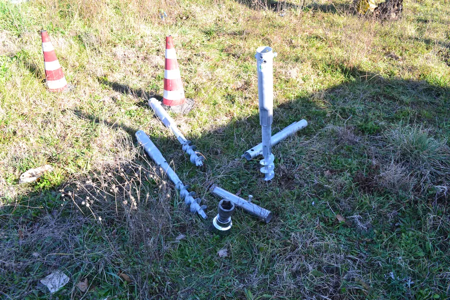 Fondazioni a vite T-Block e componenti di collegamento appoggiati sull'erba a bordo strada, pronti per la posa, accanto a un cono di segnalazione.
