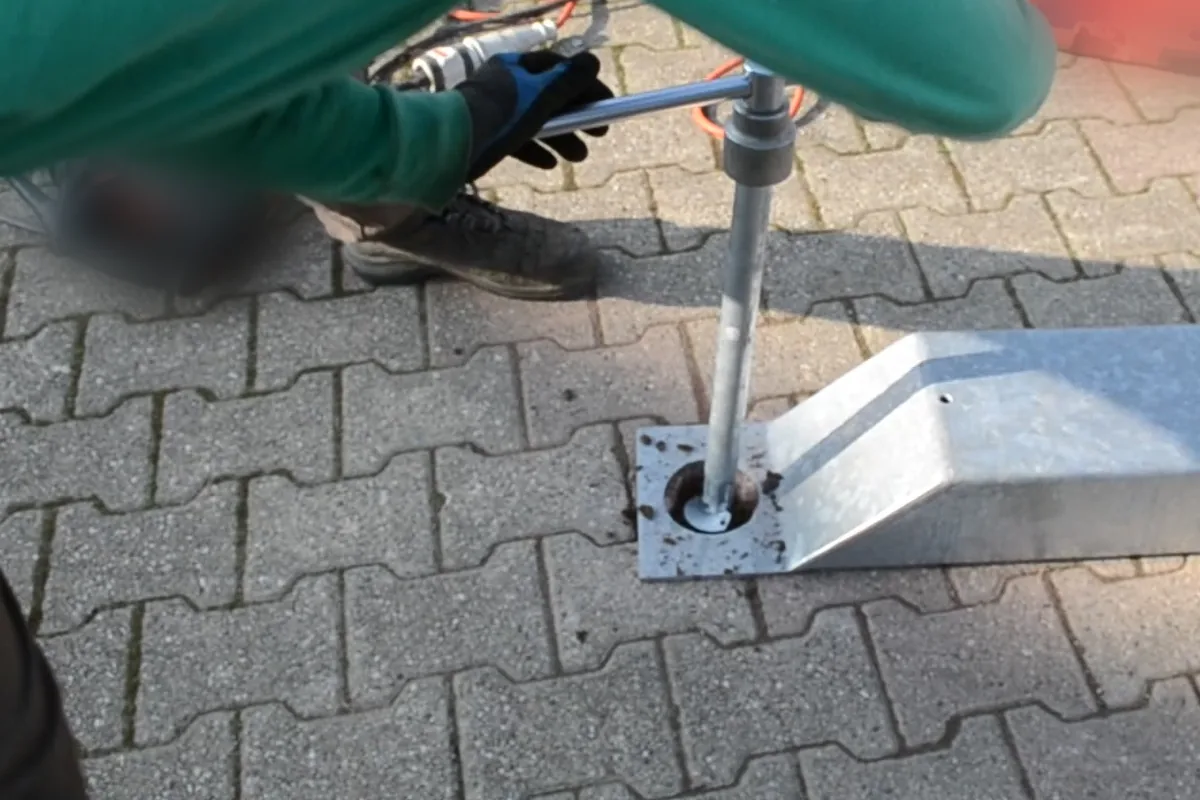 Mani con i guanti sul manico a T che avvitano un T-Block attraverso il foro della piastra di un carport, sulla pavimentazione.