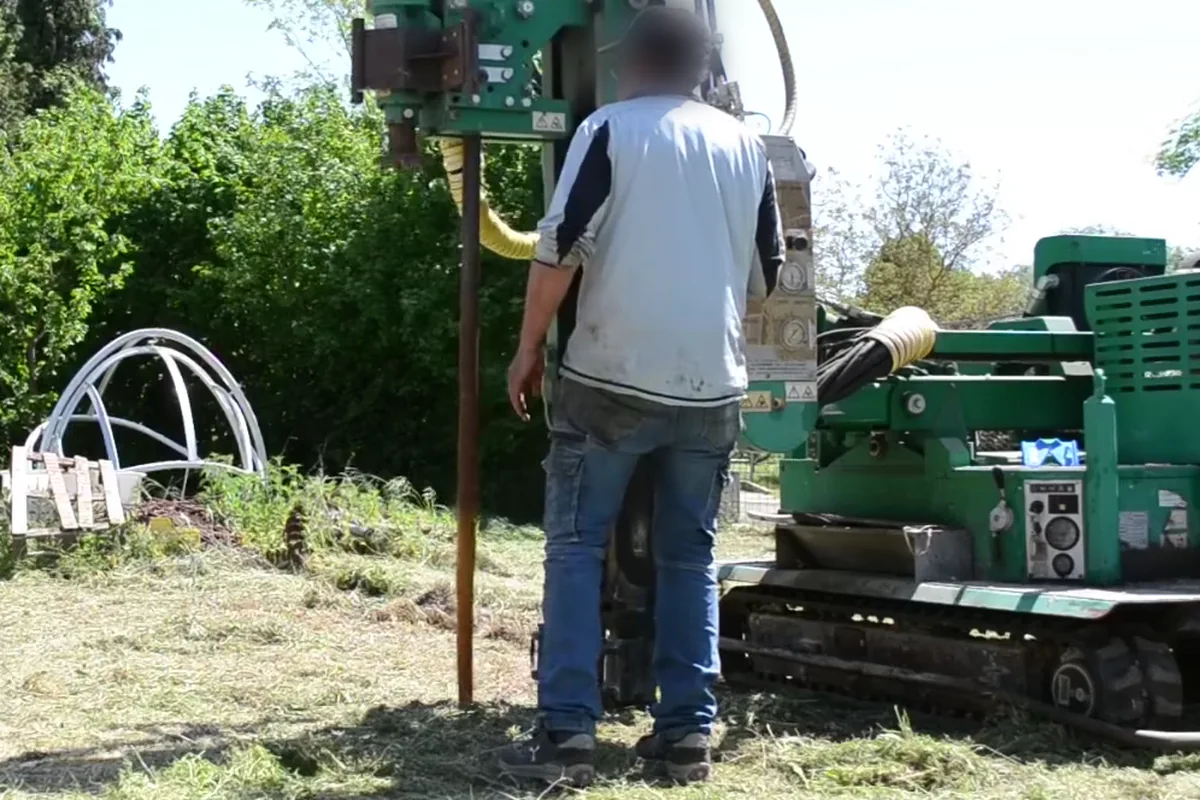 Macchina cingolata verde a colonna che apre il foro guida in un prato: l'asta da preforo scende nel terreno accanto alla colonna, con l'operatore di spalle ai comandi.