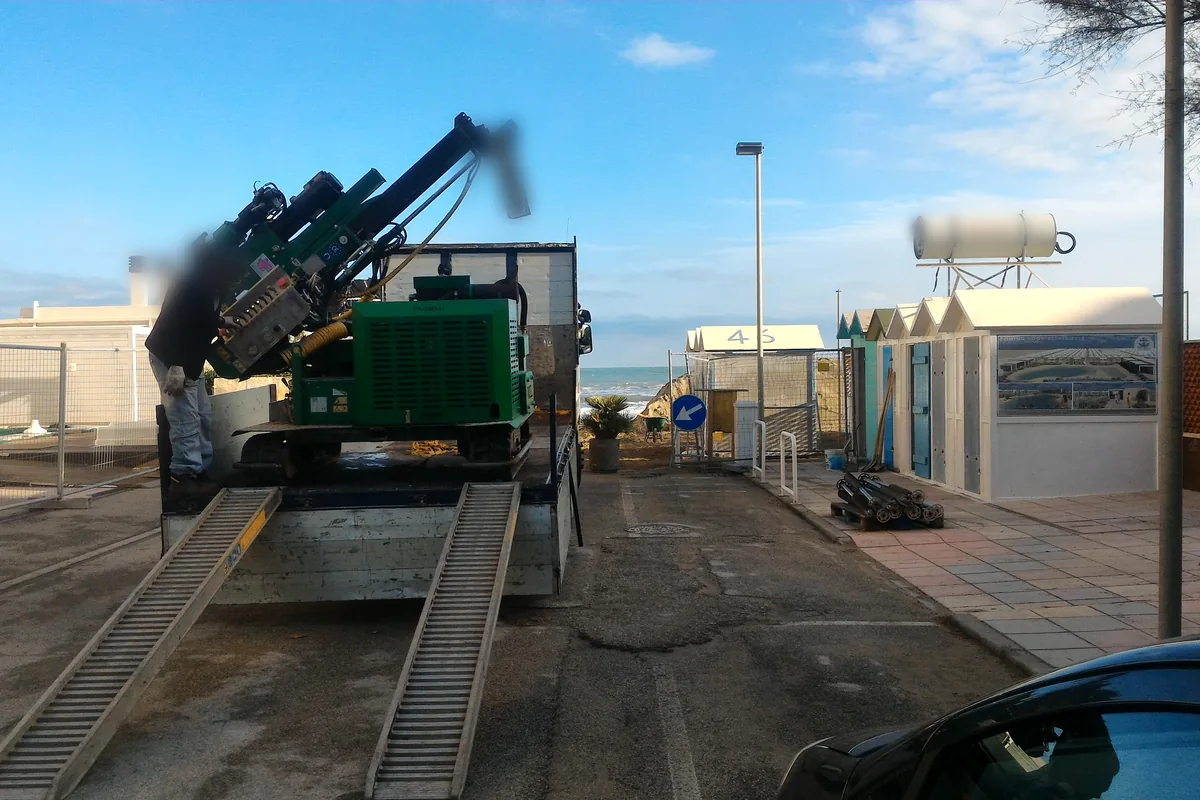 Macchina cingolata verde con la colonna ripiegata che scende dal camion sulle rampe, sul lungomare di Misano Adriatico davanti alle cabine.