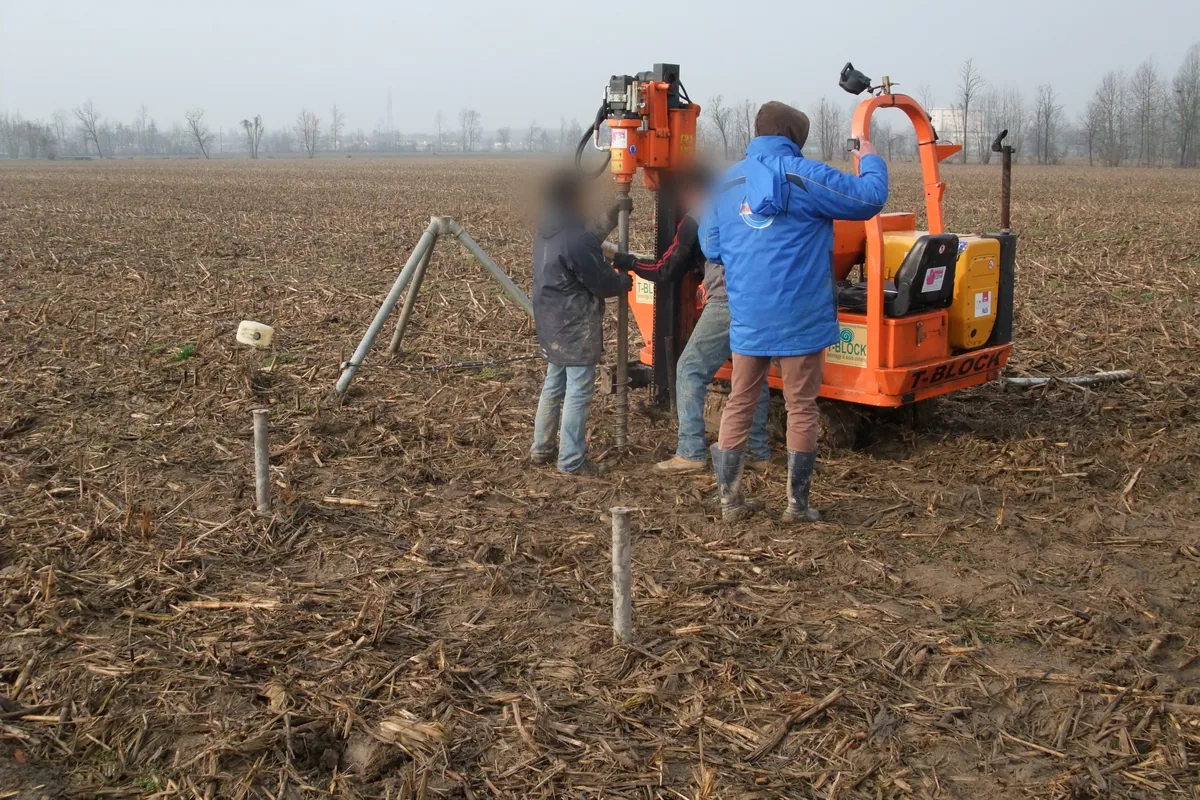 Macchina cingolata TBDRIVER arancione che avvita un T-Block nel campo arato, con il treppiede e il martinetto per la prova di un T-Block già posato in primo piano.