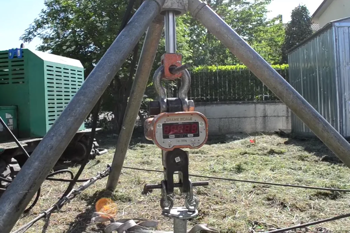 Sotto il treppiede, il dinamometro digitale appeso in linea tra lo stelo del martinetto e la testa del T-Block, in un prato accanto alla macchina cingolata.