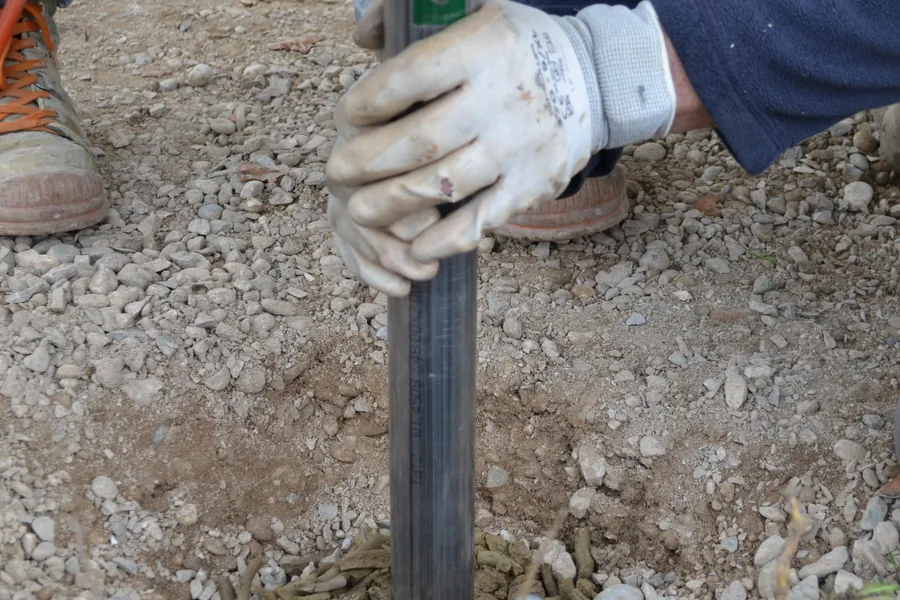 Mani con guanti da lavoro che avvitano una fondazione a vite T-Block nel terreno ghiaioso