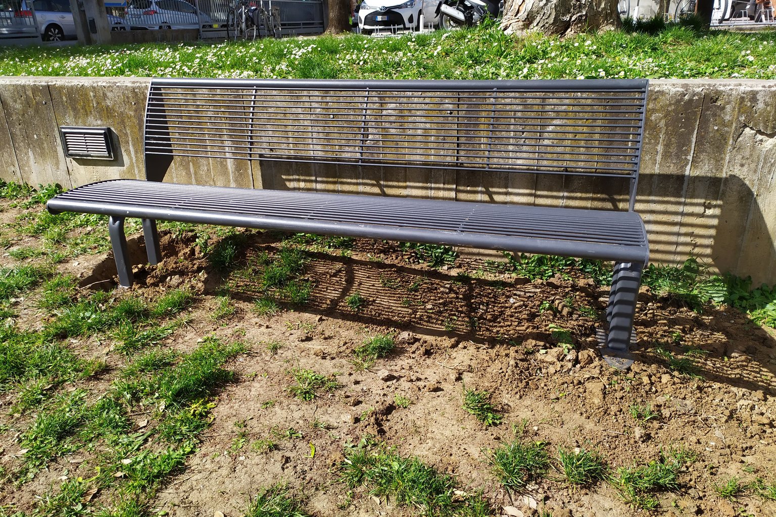 Panchina pubblica in acciaio installata in un'aiuola urbana, fissata al terreno con ancoraggi a vite T-Block, senza zavorre o calcestruzzo.