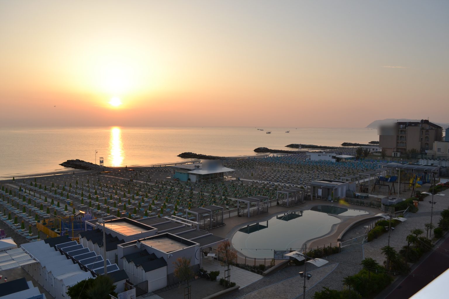 Alba sul mare a Misano Adriatico: in primo piano lo stabilimento Spiagge Luna con la piscina, le pergole bianche e le file di ombrelloni chiusi.