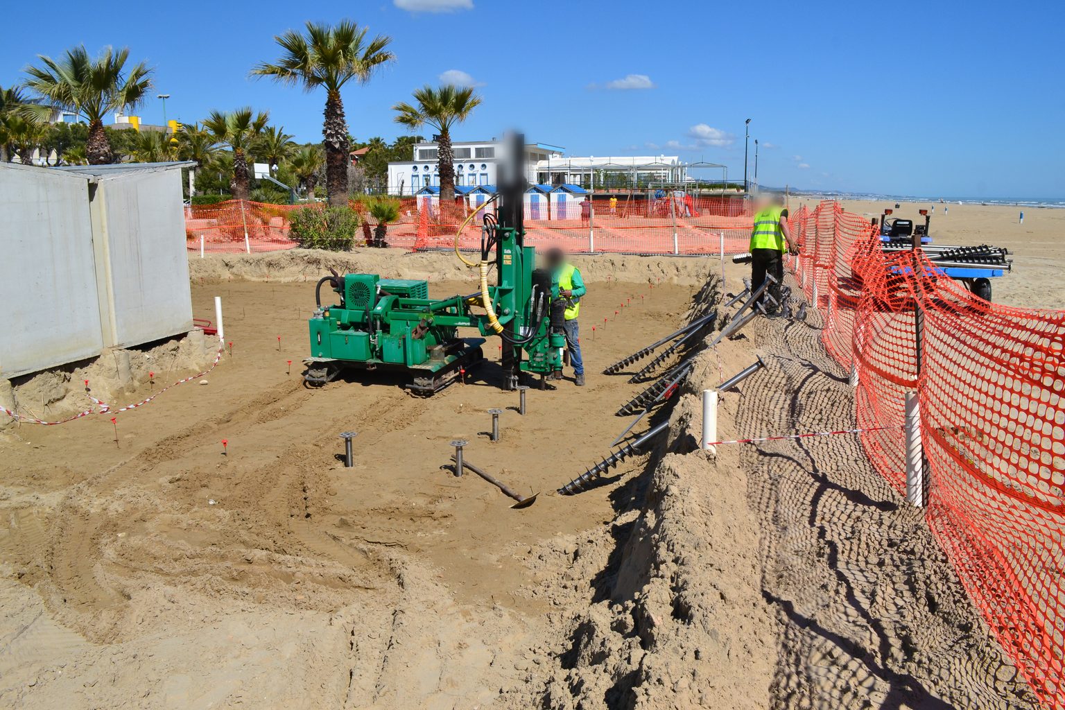 Macchina cingolata verde con la colonna verticale al lavoro sulla spiaggia di Giulianova, tra le file di T-Block già posati e i picchetti dei punti successivi.