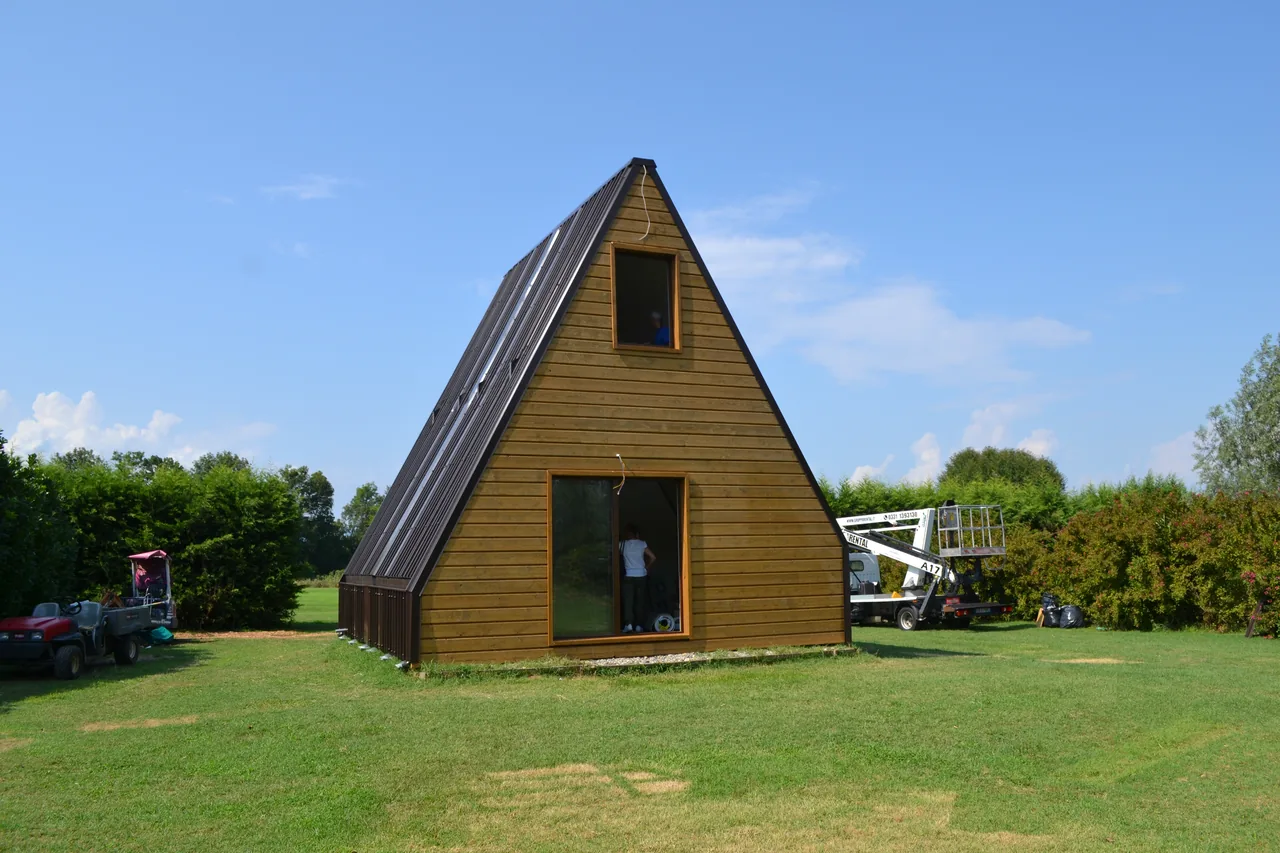 Casa modulare a forma di A con rivestimento in legno e copertura scura, montata su un prato aperto con alberi sullo sfondo.