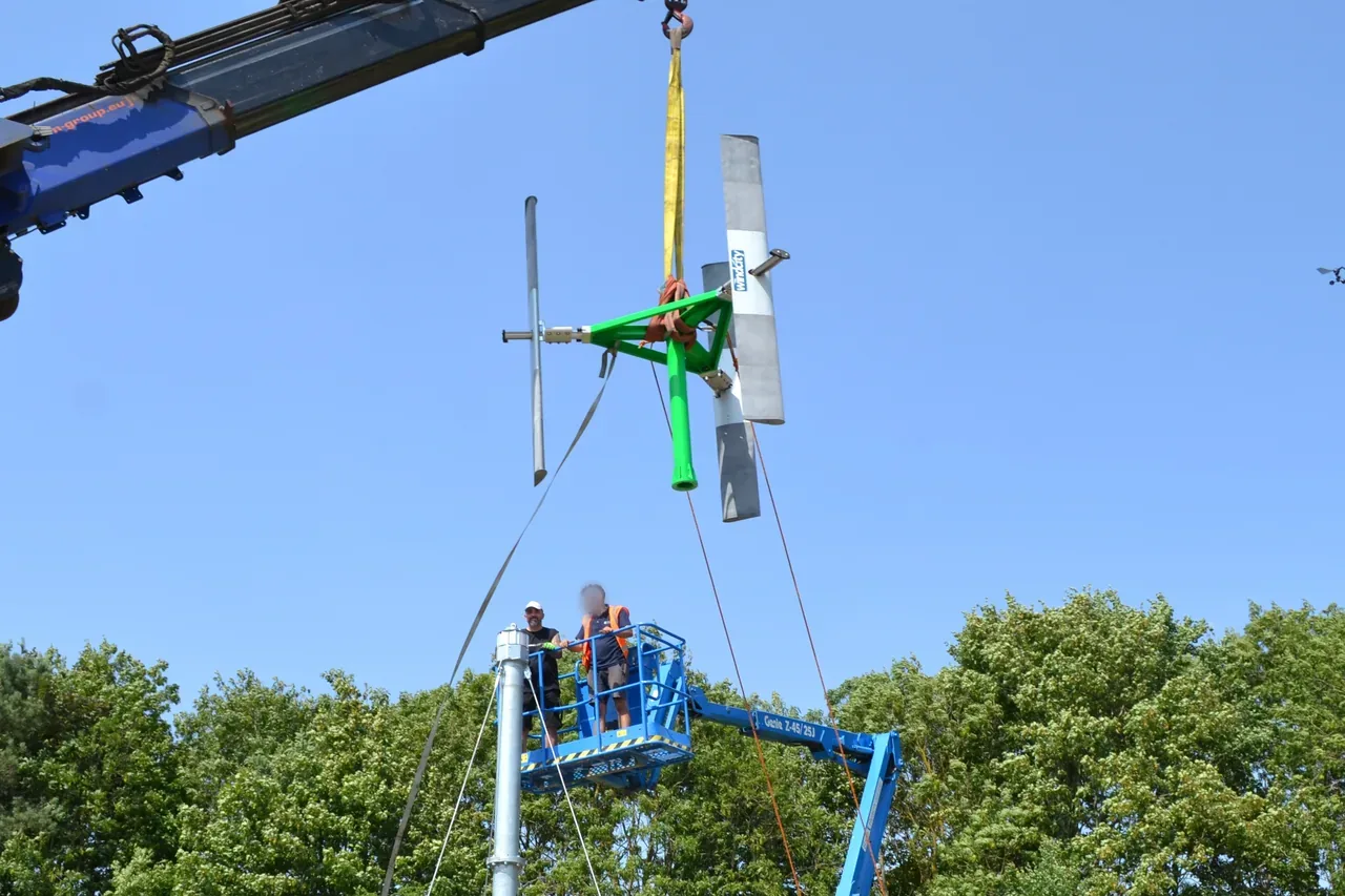 Rotore di una turbina eolica ad asse verticale sollevato da un'autogrù contro il cielo, durante il montaggio in un prato alberato.