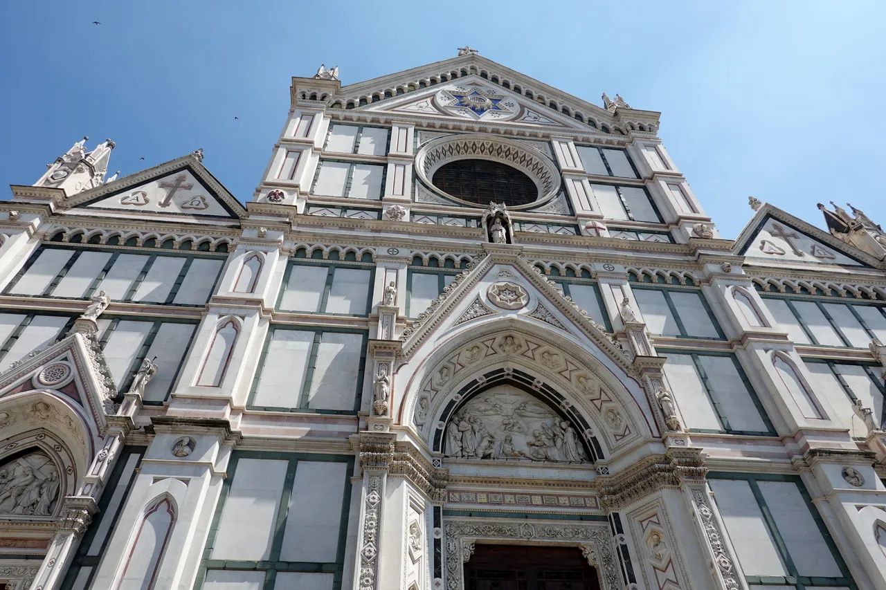 La facciata della Basilica di Santa Croce a Firenze, in marmo bianco, verde e rosa, vista dal basso contro il cielo azzurro.
