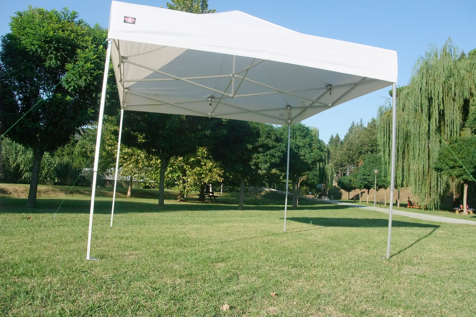 Gazebo richiudibile bianco montato sul prato di un parco, con le cime di controvento tese dal telo verso il terreno.