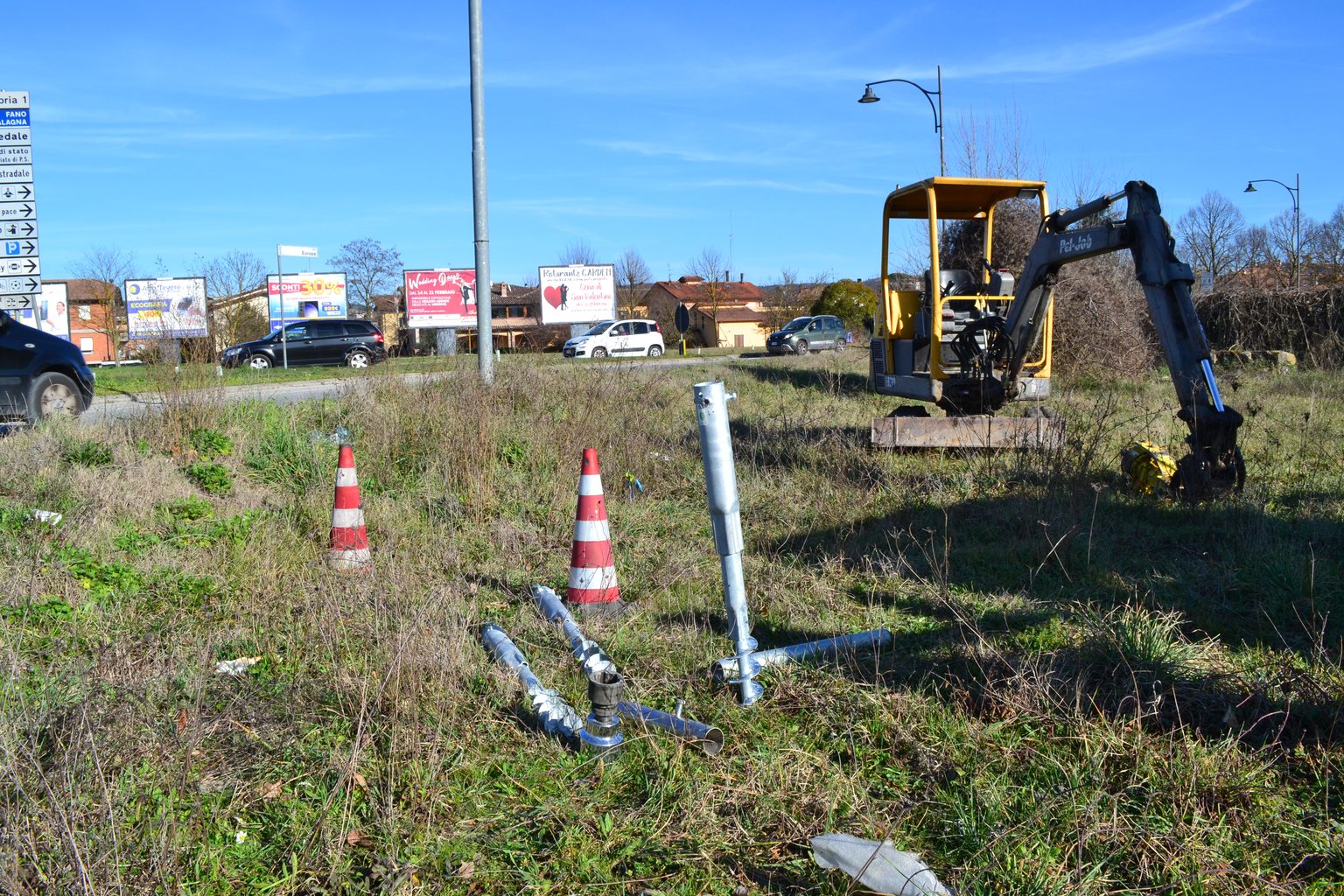 Cantiere di segnaletica stradale su banchina: fondazioni a vite T-Block in primo piano, coni di delimitazione e mini escavatore con testa idraulica per l'avvitamento.