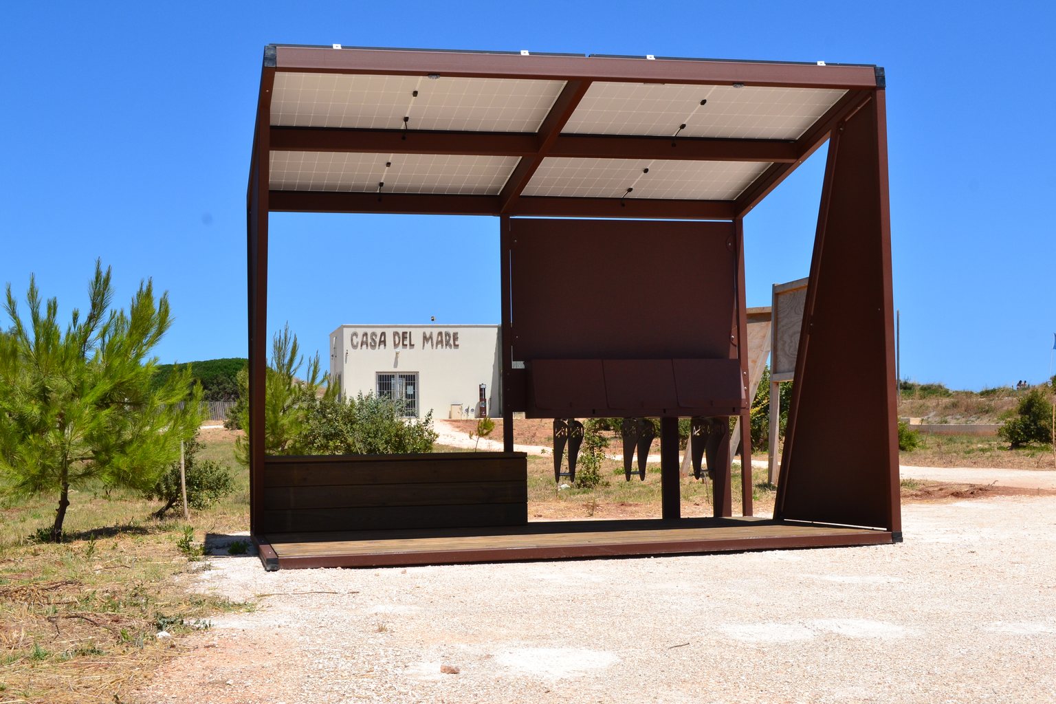 Pensilina fotovoltaica in acciaio corten con pedana in legno, su fondazioni a vite T-Block, in un parco affacciato sulla macchia mediterranea.