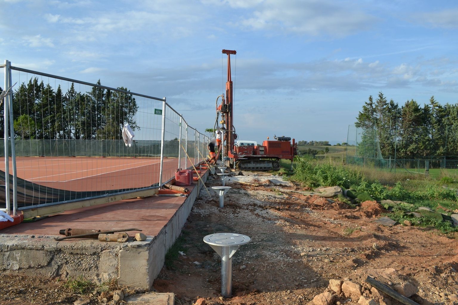 Bordo di un campo da tennis in terra rossa: una fondazione a vite T-Block già posata in primo piano e la trivella cingolata al lavoro lungo la linea successiva.