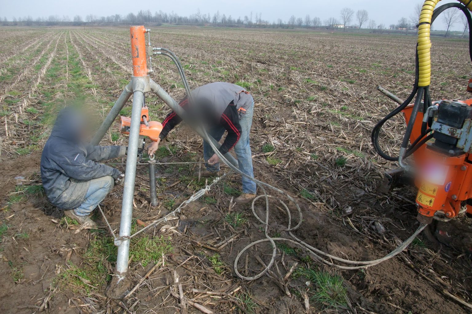 Treppiede in acciaio con il martinetto idraulico in cima collegato alla testa di un T-Block in un campo agricolo, con due operatori e la macchina cingolata.
