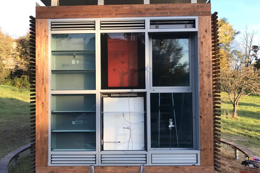 Test cell di ricerca dell'Università di Firenze in un prato: modulo rivestito in listelli di legno con vetrate e griglie di ventilazione, sollevato dal terreno sulle fondazioni a vite T-Block.