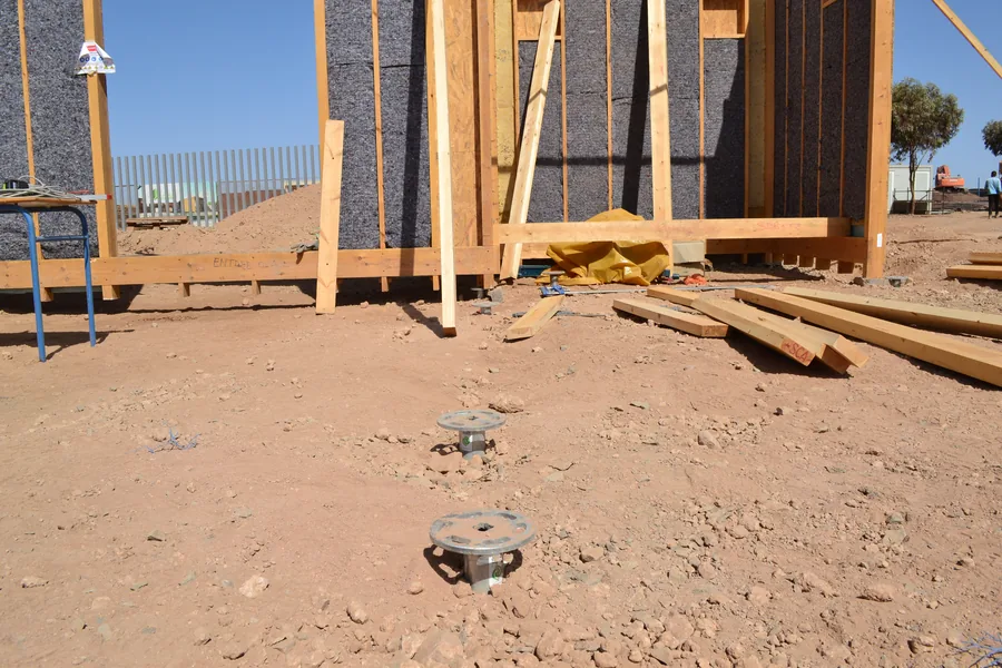 Solar Decathlon Africa a Benguerir: due teste flangiate di fondazioni a vite T-Block nella terra rossa, dietro le pareti in legno del prototipo.