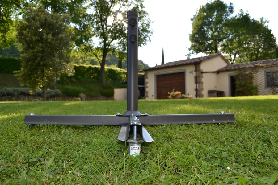 Base a croce di un grande ombrellone da giardino appoggiata sul prato: al centro si vede la fondazione a vite T-Block che la tiene ferma, al posto della zavorra in calcestruzzo.