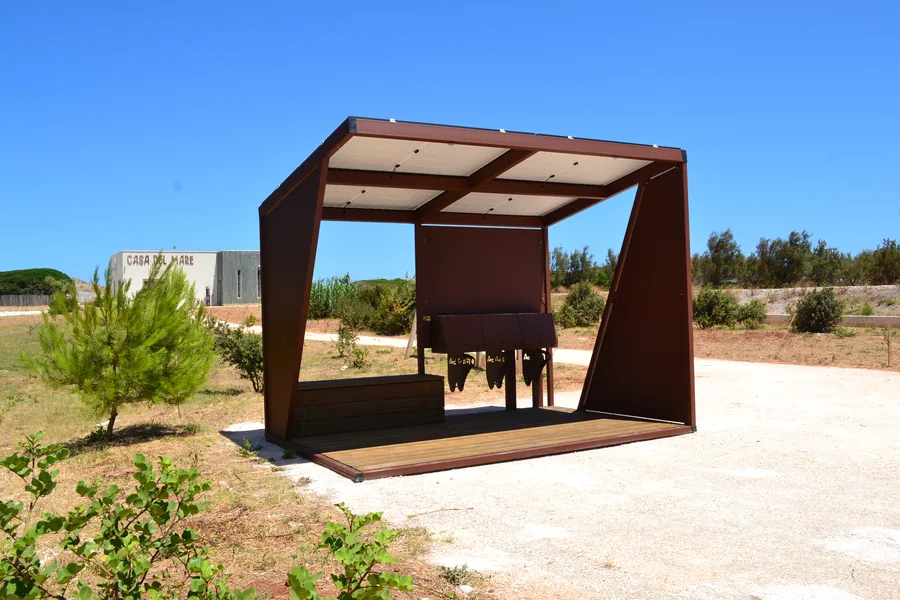 Pensilina in acciaio corten con copertura fotovoltaica, pedana in legno e sedute, in un parco pubblico affacciato sulla macchia mediterranea.