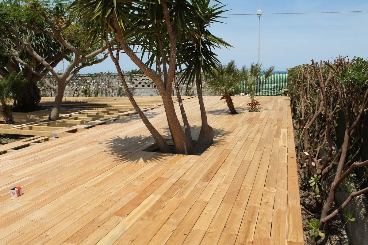 La pedana in legno finita: il tavolato chiaro gira attorno ai tronchi di una dracena e di alcune palme, con il mare in fondo oltre la siepe.
