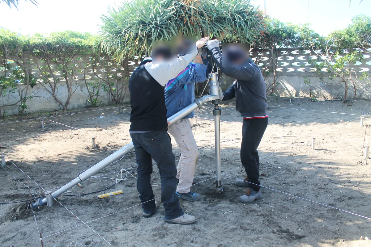 Installazione con avvitatore a barra di contrasto di una fondazione a vite nel giardino del lido: tre persone guidano l'attrezzo di avvitamento con la barra appoggiata a terra, tra i fili del tracciamento.