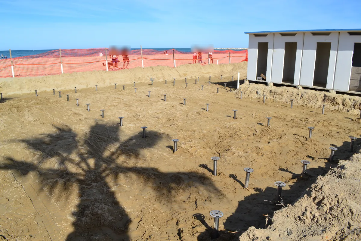 Spiaggia di Giulianova: le teste dei T-Block spuntano dalla sabbia in file regolari, attraversate dall'ombra di una palma, accanto a un blocco di cabine bianche e con il mare oltre la recinzione.