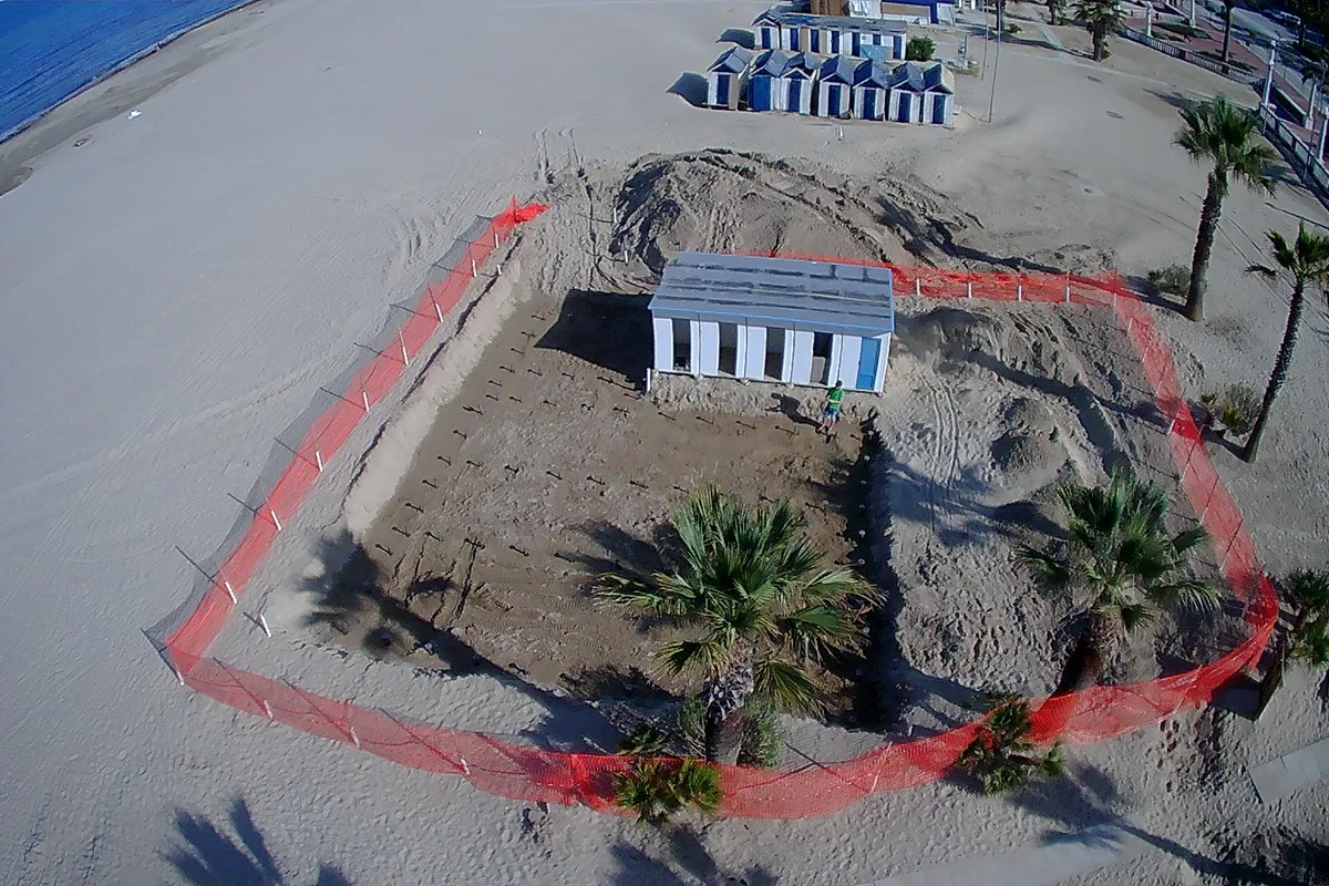 Vista dal drone: l'area di cantiere recintata sulla spiaggia di Giulianova, con il blocco di cabine, le palme e le file di fondazioni nella sabbia; in alto il mare e le altre cabine.