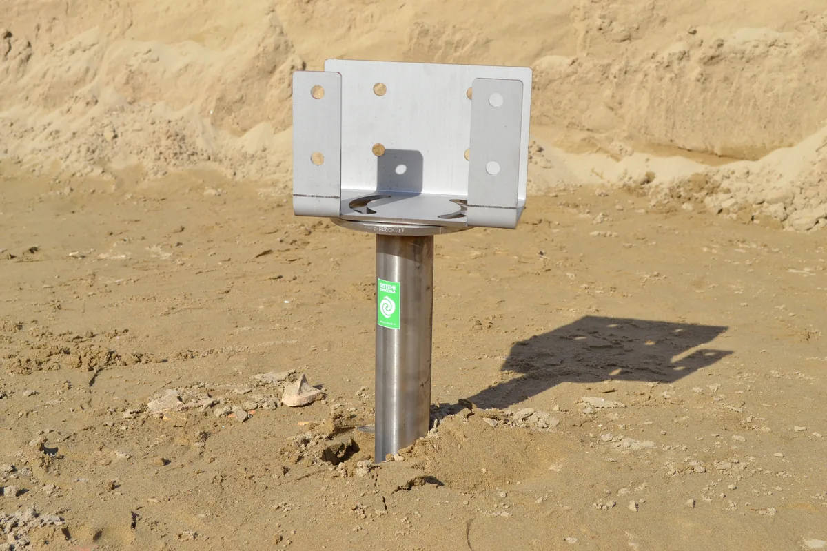 Primo piano di un T-Block in acciaio inox nella sabbia con la staffa a U fissata sulla flangia, pronta a ricevere la trave in legno.