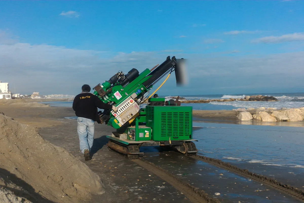 La macchina cingolata a colonna avanza sulla battigia di Misano Adriatico con un operatore di spalle accanto, il mare mosso e le scogliere sullo sfondo.