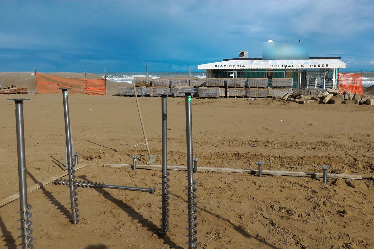 T-Block in acciaio inox appena avvitati nella sabbia di Misano, con le eliche in vista su quelli ancora da posare e il mare mosso sullo sfondo.