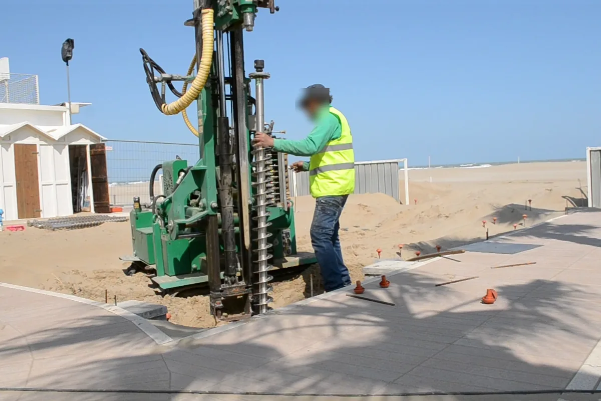 Spiaggia di Riccione: la macchina cingolata a colonna avvita un T-Block in acciaio inox nella sabbia accanto alla passerella pavimentata, con le cabine bianche a sinistra e il mare sullo sfondo.