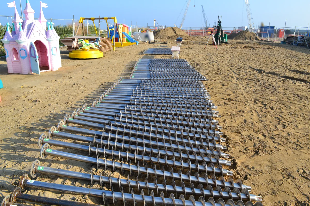File di T-Block in acciaio inox distesi sulla sabbia di Rimini prima della posa, con le eliche e le flange in vista e i giochi del parco bimbi sullo sfondo.