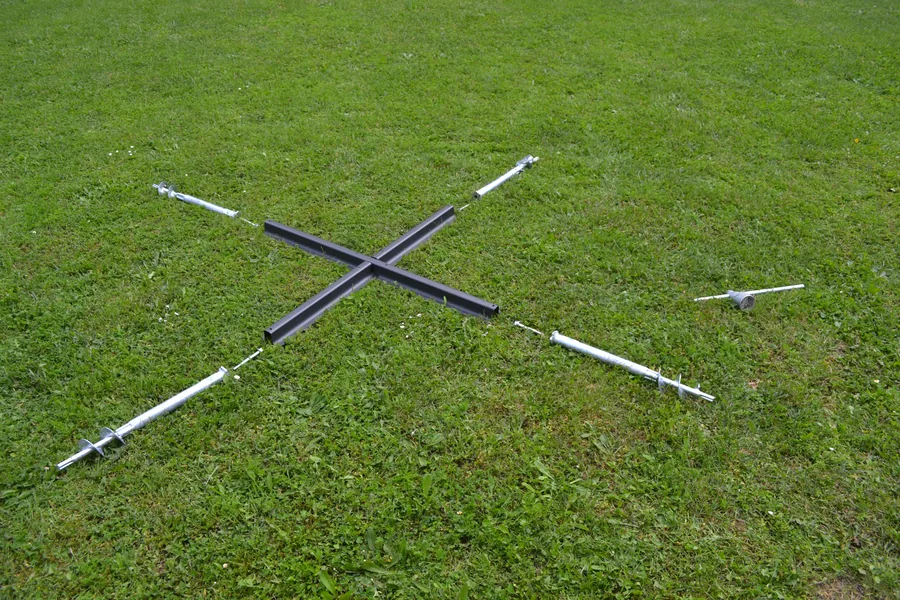 La croce di base di un ombrellone appoggiata sul prato con le quattro viti accanto: serve da dima per segnare dove andranno.