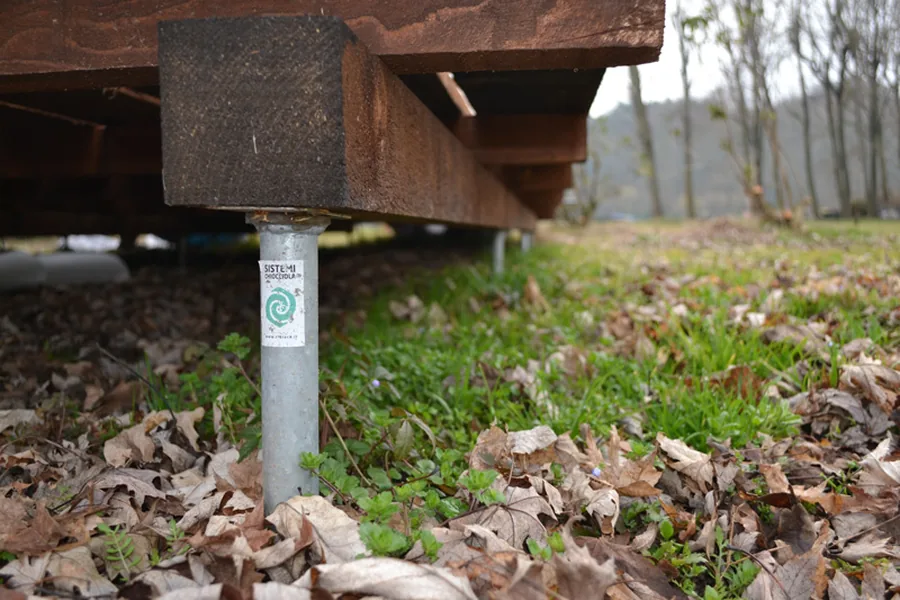 Dettaglio di una trave in legno della pedana appoggiata e bullonata sulla testa di una fondazione a vite T-Block, con il terreno e le foglie intatti tutt'intorno.