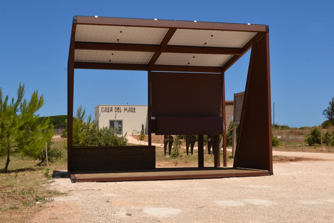 Pensilina fotovoltaica per la ricarica delle biciclette con copertura in pannelli solari, installata su fondazioni a vite T-Block in un parco pubblico vicino al mare.