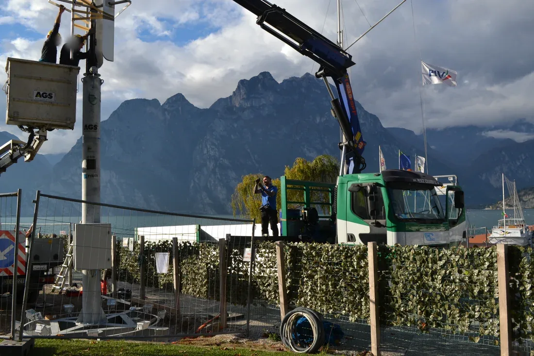 Autogrù che solleva il palo del micro-aerogeneratore, con il lago e le montagne sullo sfondo.