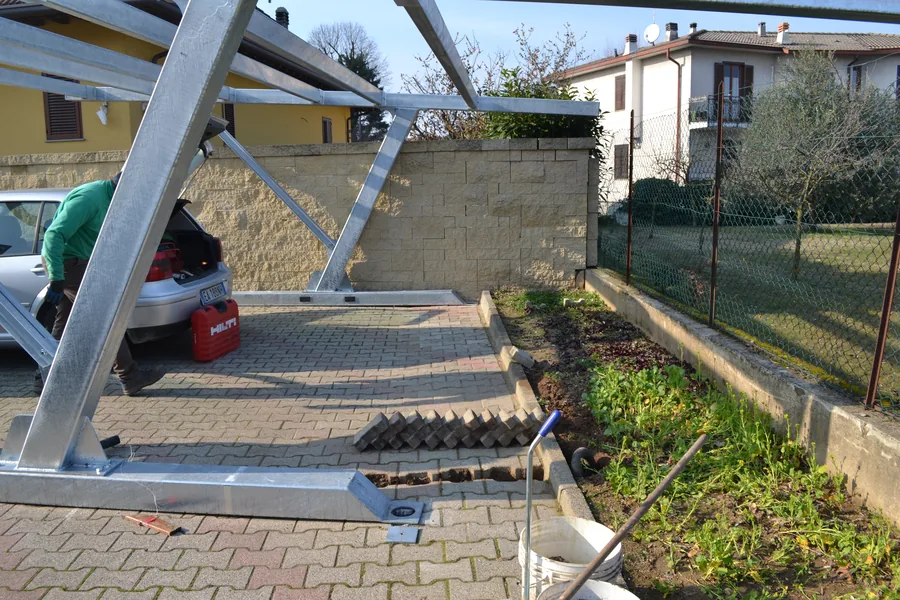Struttura di un carport in acciaio in corso di montaggio sopra un piazzale gia' pavimentato, con l'auto parcheggiata di fianco: si lavora per file, senza chiudere il parcheggio.