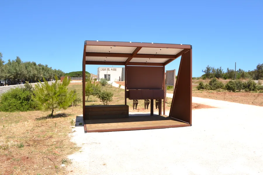 Pensilina in acciaio corten con copertura fotovoltaica e pedana in legno, installata in un parco pubblico per la sosta e la ricarica delle biciclette.