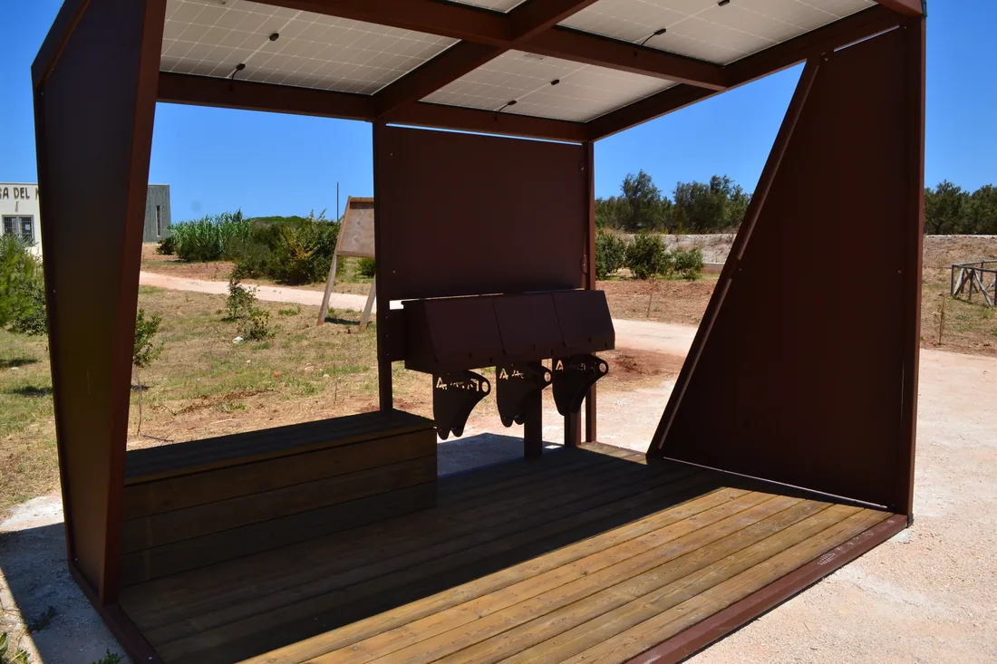 Interno di una pensilina fotovoltaica in acciaio corten con pedana in legno e sedute, in un parco pubblico.
