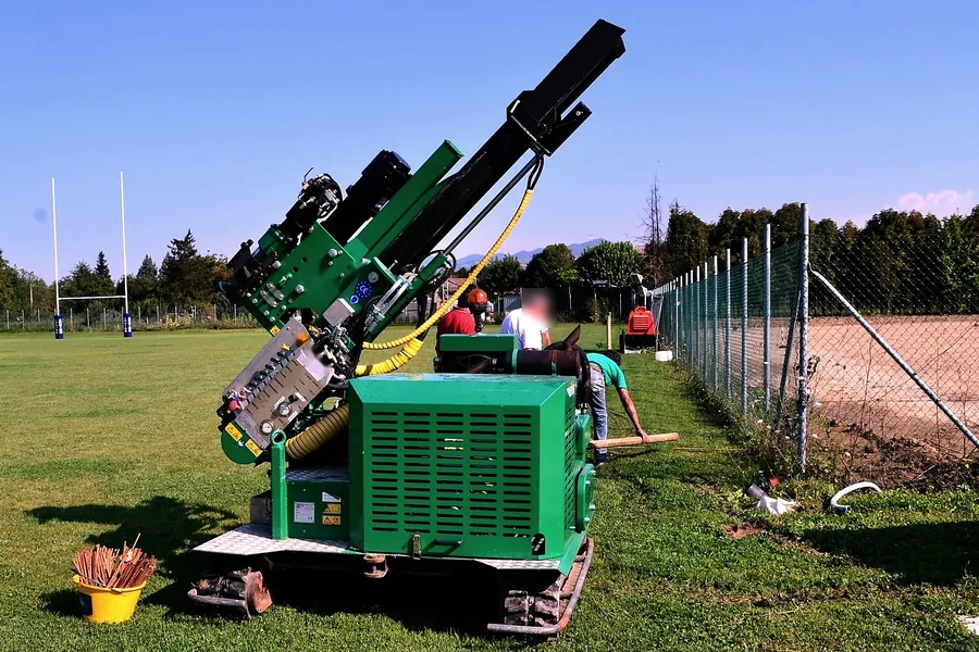 Macchina cingolata verde a colonna per l'avvitamento dei T-Block, ferma sul prato di un campo sportivo.