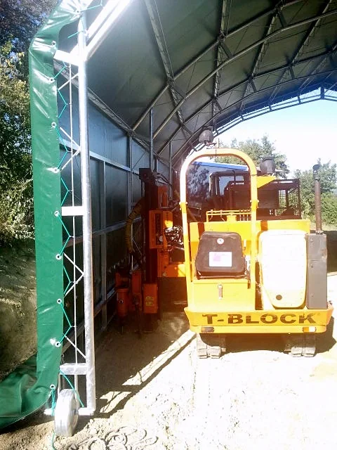 Tunnel agricolo con telo verde e, sotto la copertura, la macchina cingolata arancione T-Block per l'avvitamento delle fondazioni a vite.