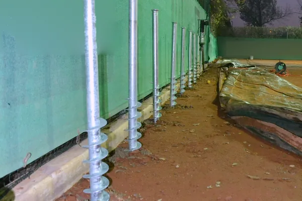 Fila di fondazioni a vite T-Block lungo il bordo di un campo da tennis in terra rossa, per l'ancoraggio della copertura.