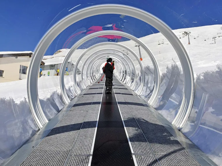 Tapis roulant di risalita coperto da un tunnel trasparente ad archi in un comprensorio sciistico innevato, con uno sciatore che lo percorre e le piste sullo sfondo.