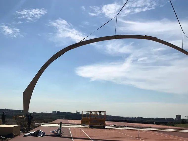 Grande arco in legno lamellare montato sopra i campi da tennis, stagliato contro il cielo, sostenuto alle estremita' dalle fondazioni a vite T-Block.