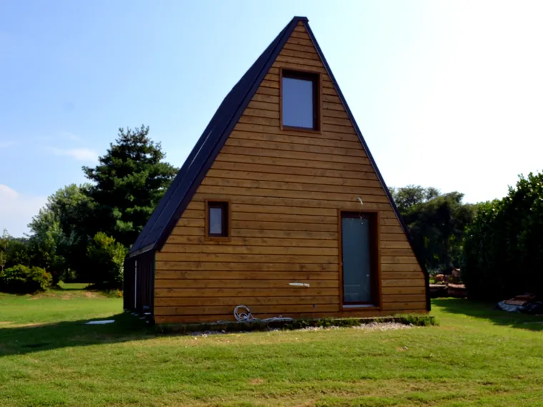 La casa pieghevole finita, in legno a forma di A, appoggiata sul prato senza platea di calcestruzzo.